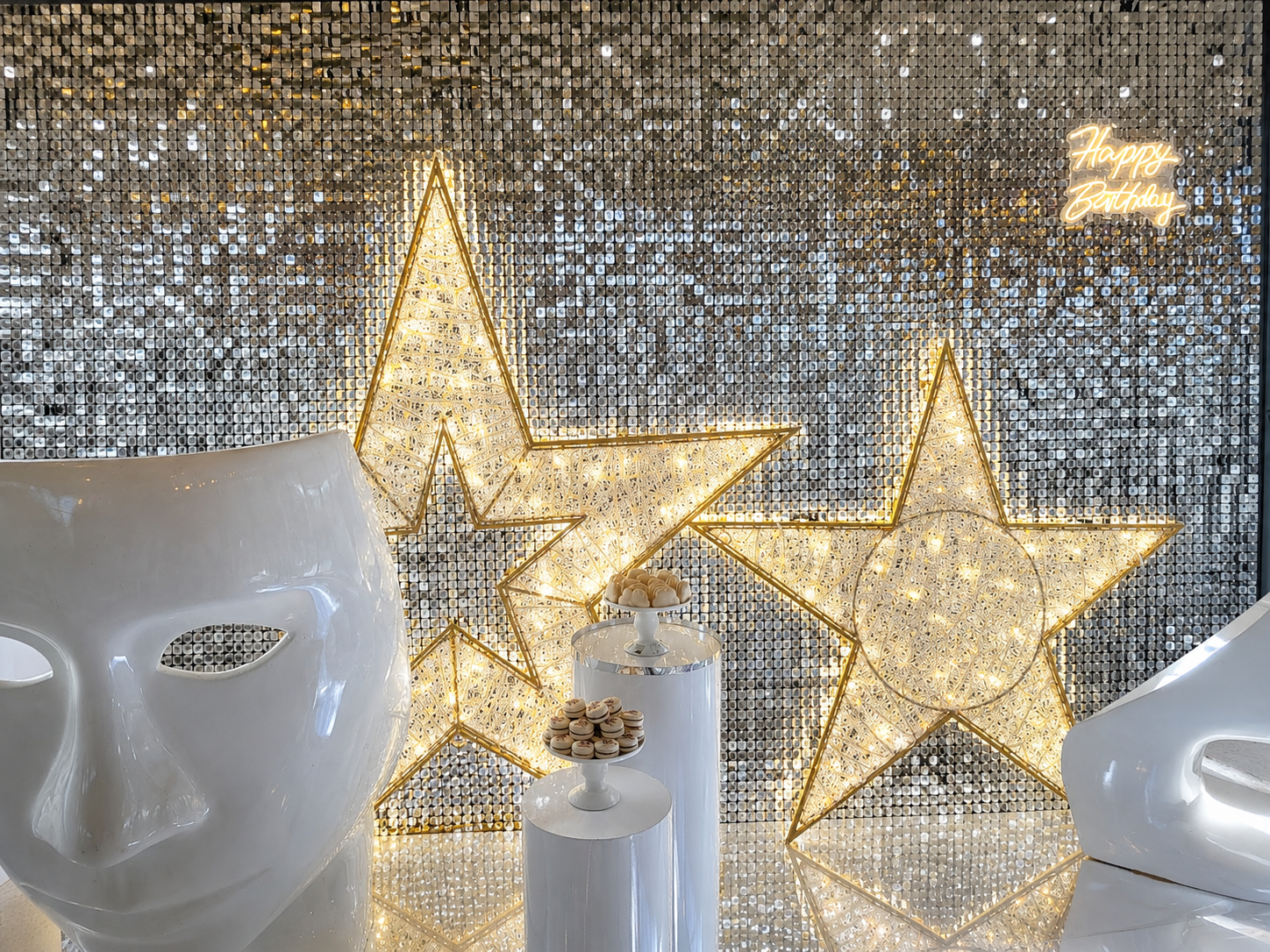 A festive holiday display featuring two large glowing star-shaped light decorations, white cylindrical candle holders with gold ornaments, and white decorative masks in front of a sparkling silver sequin backdrop with a neon 'Happy Birthday' sign.