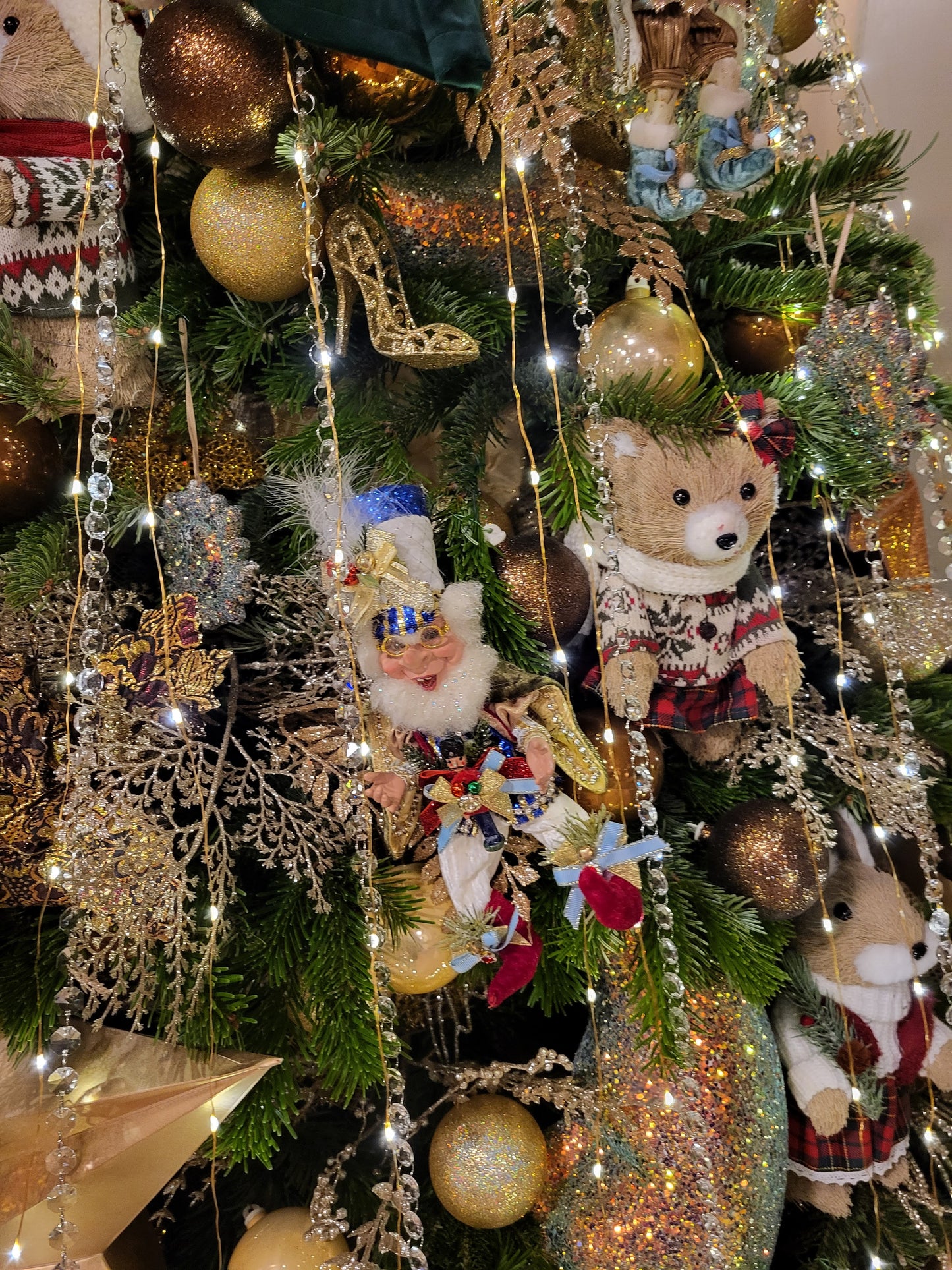 A close-up of a decorated Christmas tree with green pine branches, gold and bronze ornaments, string lights, glittery garlands, lace ornaments, and plush teddy bears dressed in holiday clothing.